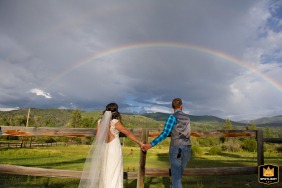 A bride and groom, at Perry Mansfield Performing Arts School in Steamboat Springs, Colorado, stand by a fence. They look at a rainbow as they stand in nature.